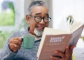 Homem idoso sorridente lê um livro enquanto toma seu café. Recomendando livros curtos para ler em um dia.