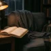 Poltrona com algumas cobertas sobre ela. Ao lado, uma mesa de canto com uma luminária antiga acesa e um livro aberto sobre ela. Ao fundo, uma janela mostrando a noite neblinada e uma estante cheia de livros. Falando sobre os melhores livros de suspense.
