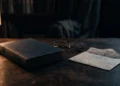 Livro antigo sobre uma mesa, junto a um óculos, uma carta semiaberta e uma luminária. O ambiente está em luz baixa. Falando sobre autores parecidos com harlan coben.